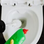 Top view crop anonymous person wearing yellow cleaning gloves cleaning toilet while pouring liquid detergent from green bottle in toilet bowl