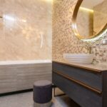 Interior of light bathroom with sink with faucet on cabinet under mirror near pouf in front of toilet next to bathtub with shower